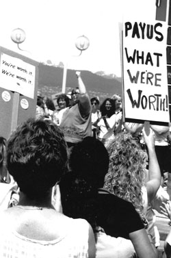 Debra McPherson with fist in the air in the background - Pay Us What We're Worth sign in foreground